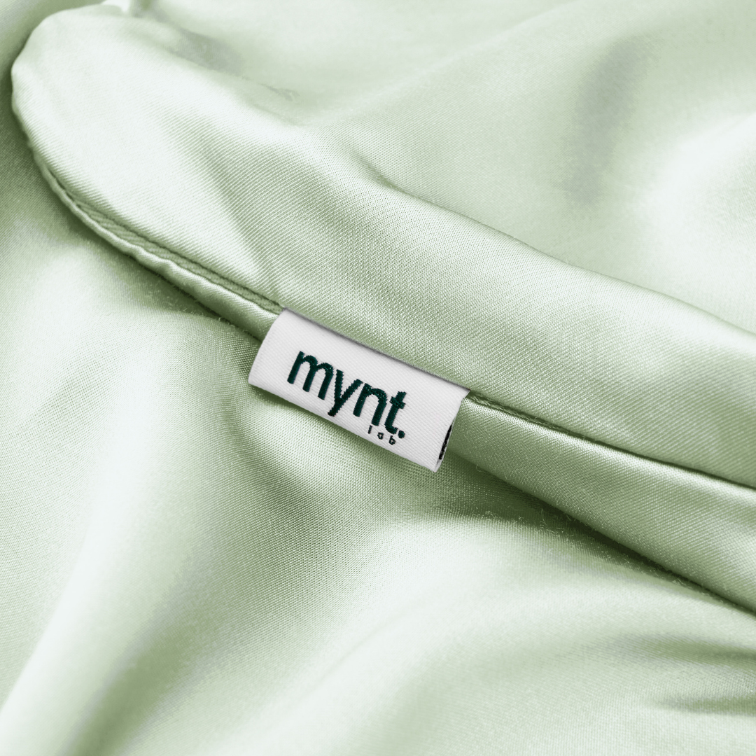A close-up of the Fitted Sheet Set in light green, featuring a white mynt lab tag stitched on. The fabric looks smooth, shiny, and silky soft.