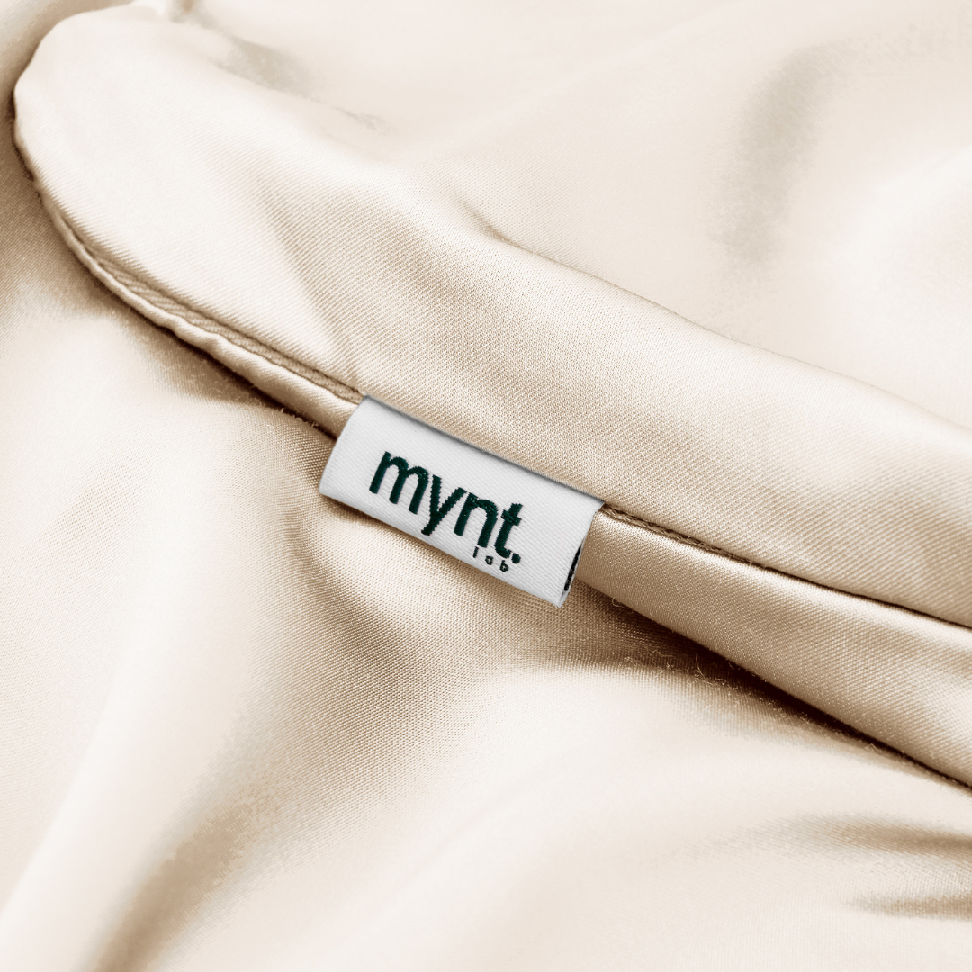 A close-up of cream satin fabric with a white "mynt" tag, partially tucked into a seam—ideal for those seeking silky soft comfort with the Fitted Sheet Set (3-Piece).