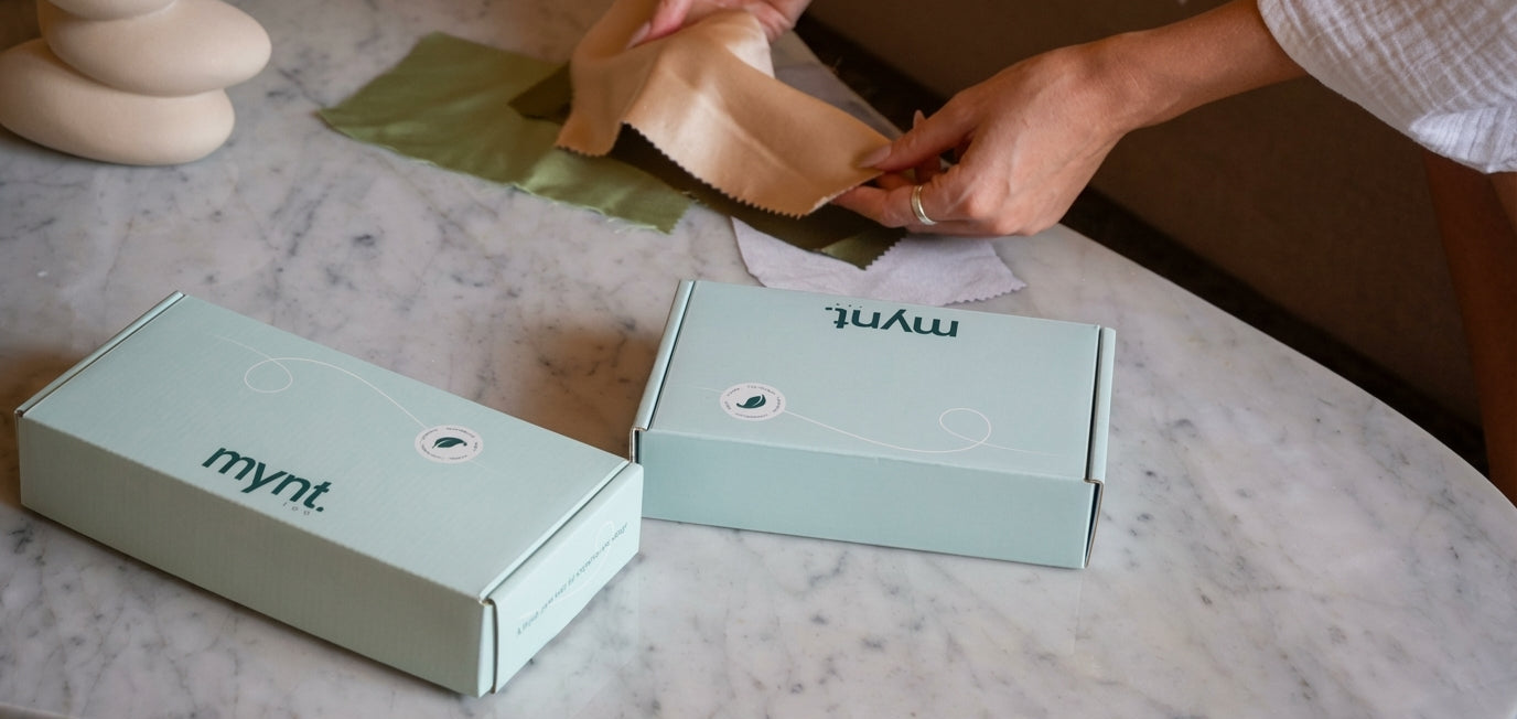 Hands holding MyntFusion fabric swatches in blush pink and sage green, with Mynt Lab packaging boxes on marble surface.