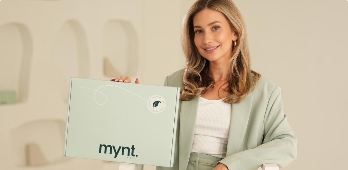 A woman with long blonde hair, wearing a light green suit and white top, sits on a chair indoors holding a light green box labeled mynt. She is smiling and facing the camera.