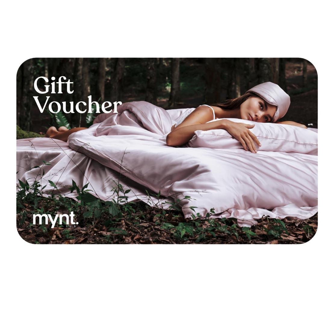 A woman in sleepwear lies on a bed with pink bedding outdoors in a forest. “Mynt Lab Gift Card” and the Mynt Lab logo are displayed on the left side of the image.