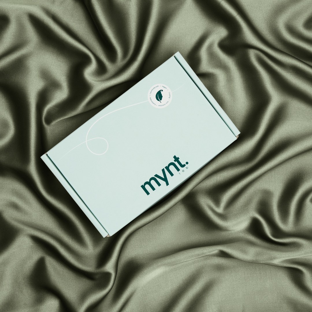 A light green rectangular box with the mynt. logo sits atop matching green satin fabric, creating a soft and elegant background.