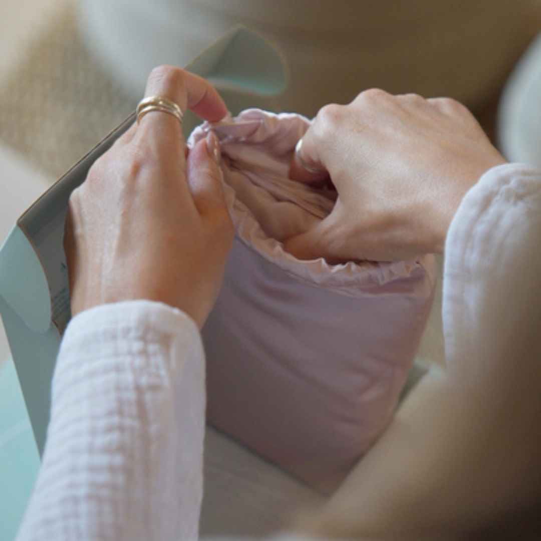 A person with light skin opens a light pink, fabric bag or pouch with both hands. The person is wearing a white long-sleeve top and gold rings. The background is softly blurred.