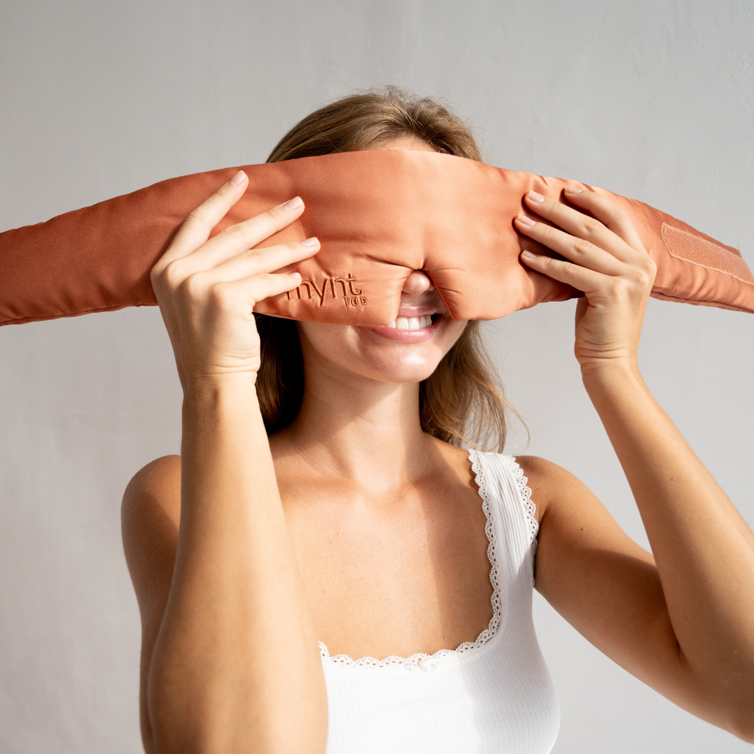 A smiling woman in a white tank top holds a rust-colored eye pillow over her eyes against a plain light background.
