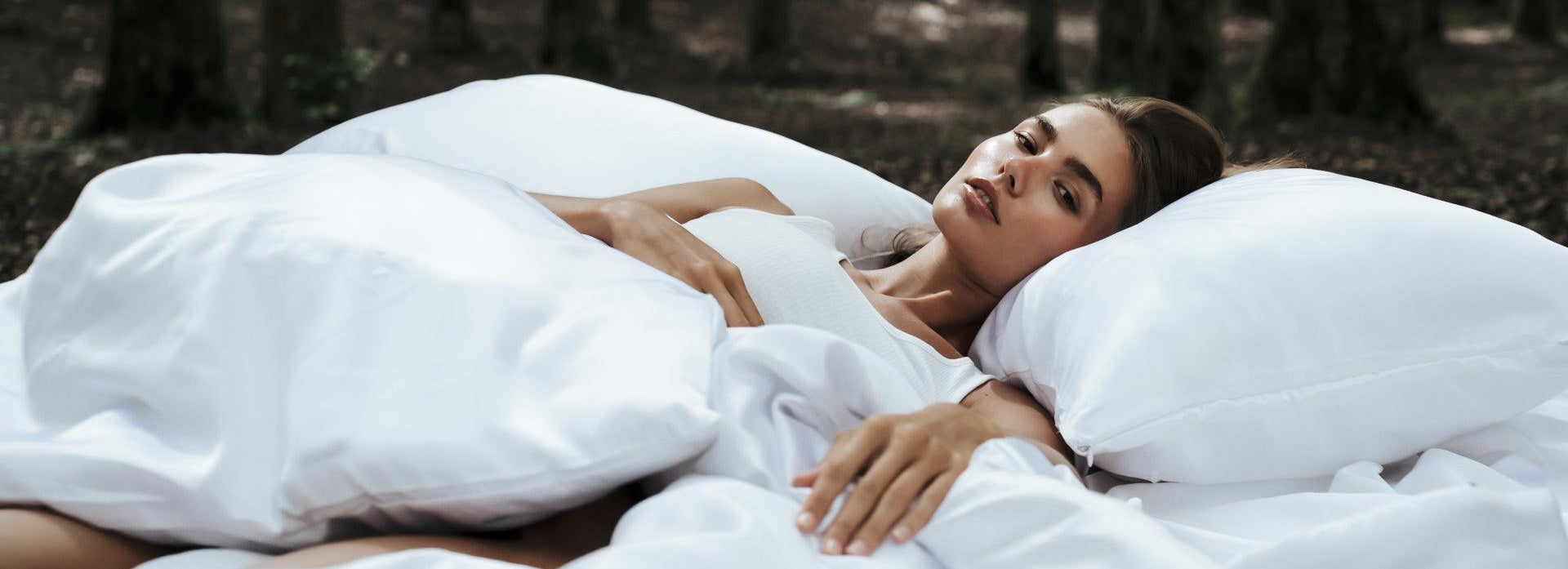 A woman lies on a bed with white sheets and pillows in the middle of a forest, looking relaxed and contemplative. Sunlight filters through the trees in the background.