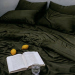 An open book, two lemons, and a glass of water sit on dark green MyntFusion™ Complete Bedding Bundle by Mynt Lab, with matching pillows visible in the background of an unmade bed.