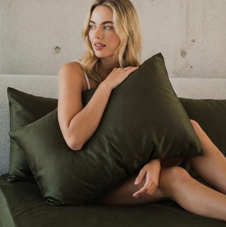 A blonde woman sits on a sofa, holding a large dark green pillow and relaxing—just as she would on the ultra-soft Mynt Lab MyntFusion™ Fitted Sheet Set (3-Piece).