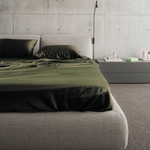 A modern bedroom with a low, upholstered bed featuring dark green sheets and pillows, next to a minimalist gray nightstand holding small items, all set against a concrete wall.