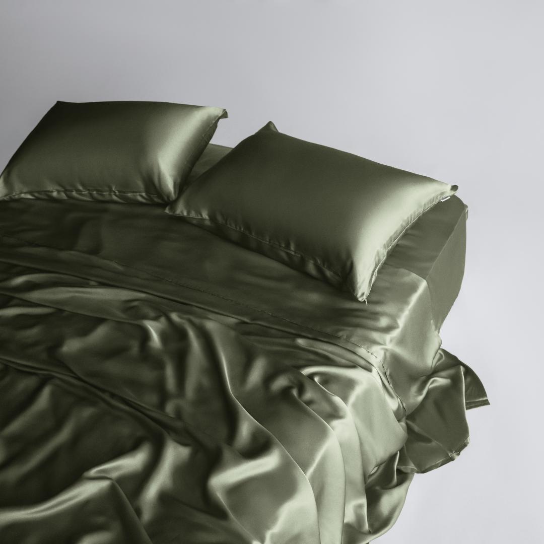 A made bed with Mynt Lab’s MyntFusion™ Complete Sheet Set (4-Piece) in olive green satin, including two matching pillows, is seen from above on a light background. The silky, cooling sheets appear shiny and drape softly off the sides.
