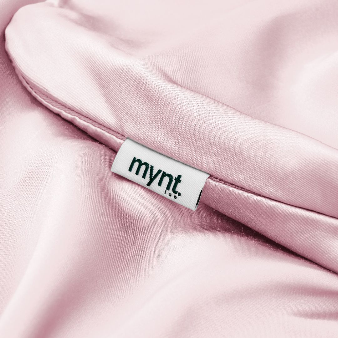 A close-up of soft, light pink silky fabric with a white Mynt Lab tag. This smooth, shiny Pillowcase helps reduce frizz and supports better beauty sleep.