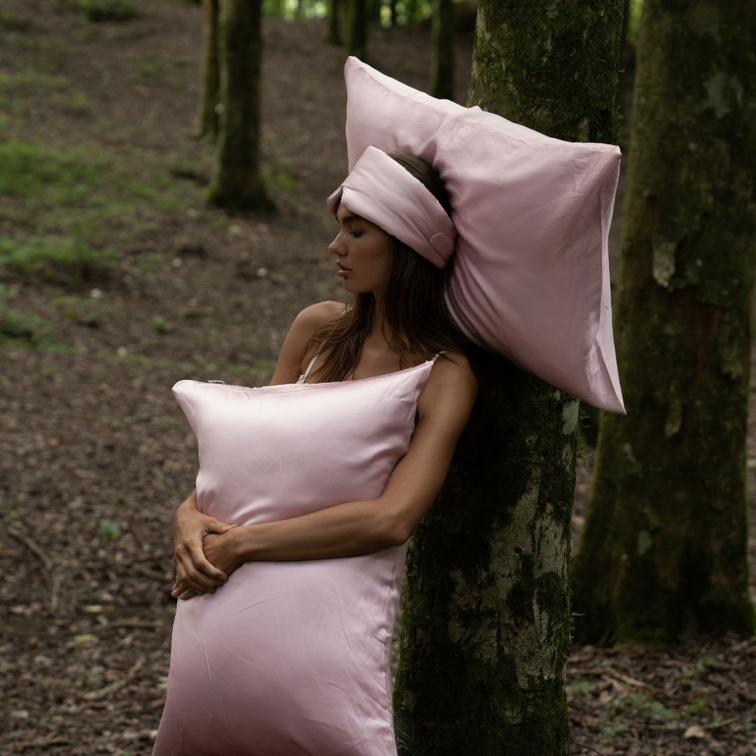 A woman in a pink sleep mask leans against a tree, resting on Mynt Lab's Pillowcase. She looks calm and relaxed, enjoying beauty sleep that reduces frizz and keeps her skin hydrated.