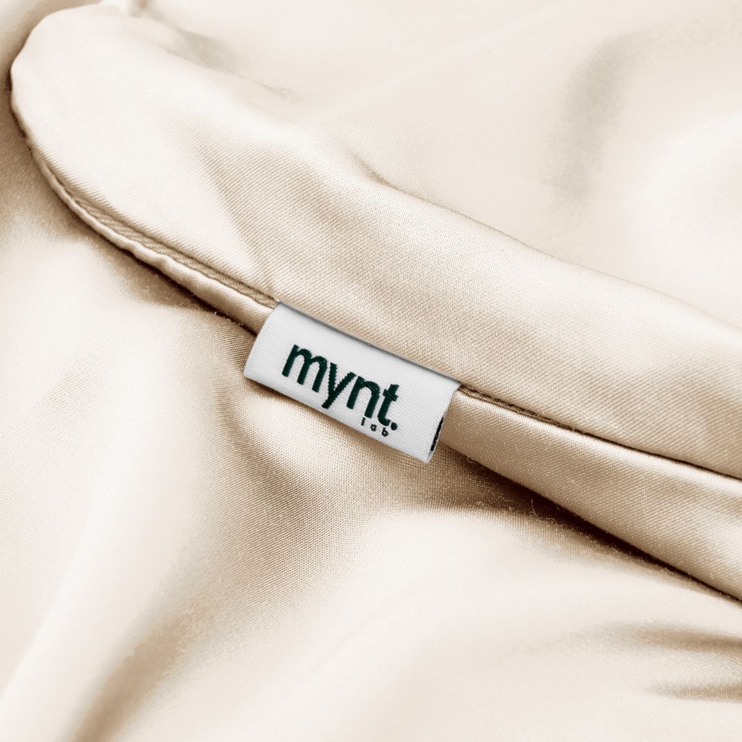 A close-up of a sand silky Pillowcase from Mynt Lab, featuring a white "mynt lab" tag. Its smooth, shiny texture helps reduce frizz and maintain hydrated skin - ideal for beauty sleep.