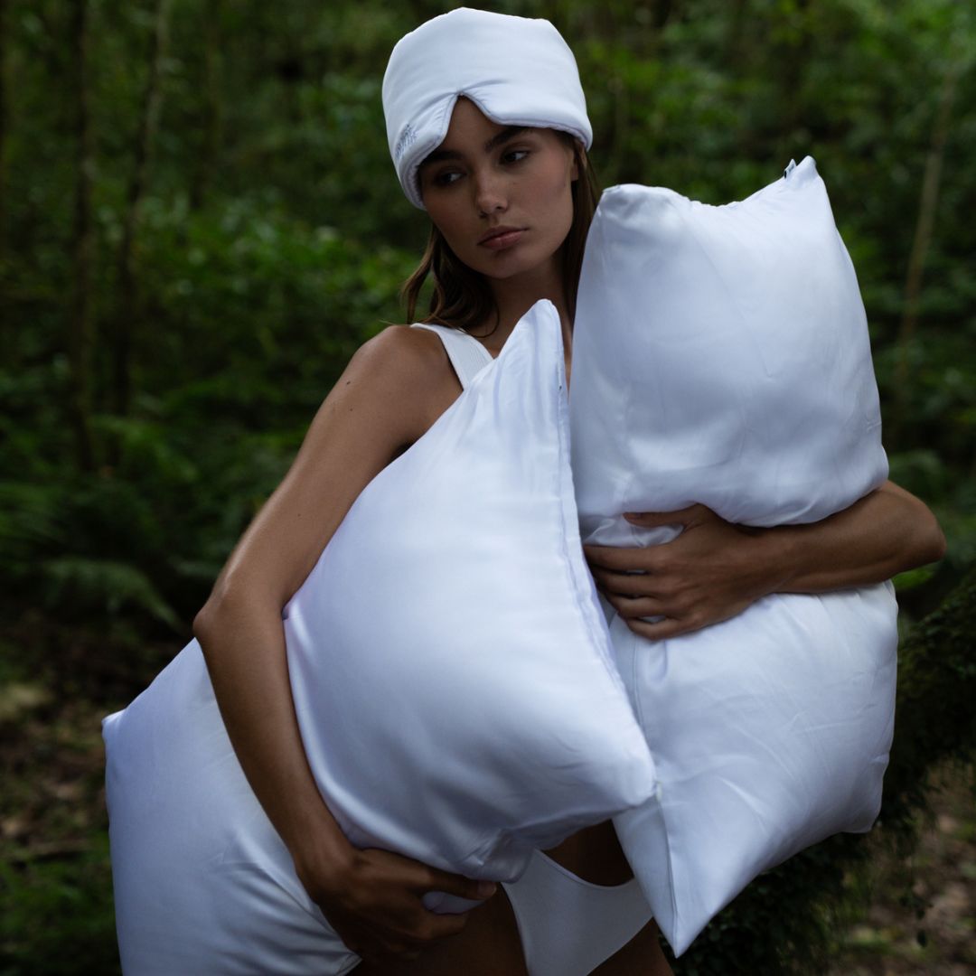 A woman in white lingerie and sleep mask stands in a lush forest, holding two large pillows, dreaming of beauty sleep and hydrated skin as she gazes thoughtfully into the distance.