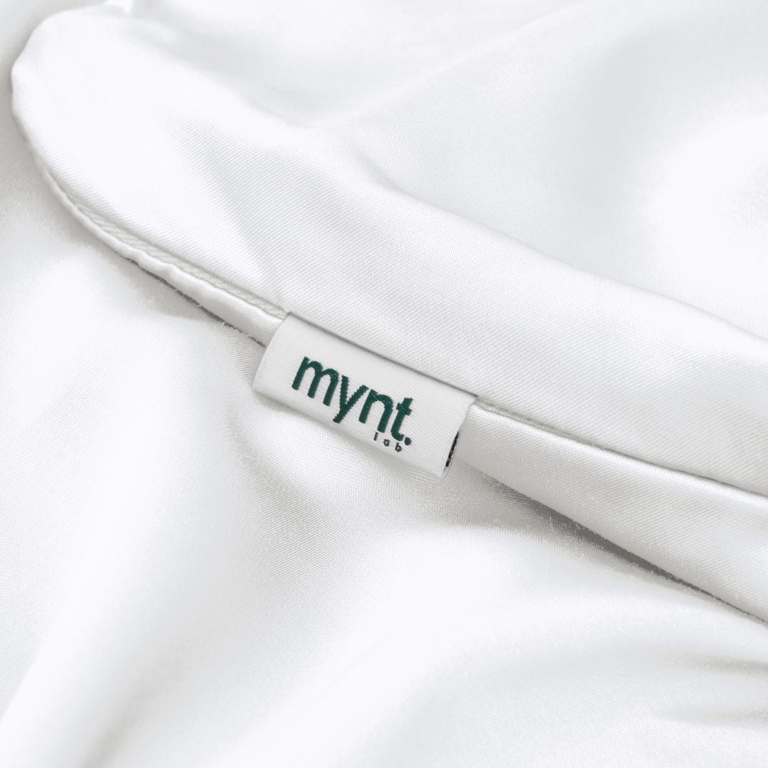 A close-up of a white Mynt pillowcase shows a sewn-in tag with "mynt lab" in dark green letters. The smooth, silky fabric is ideal for beauty sleep and may help reduce frizz, making it perfect for bedding.