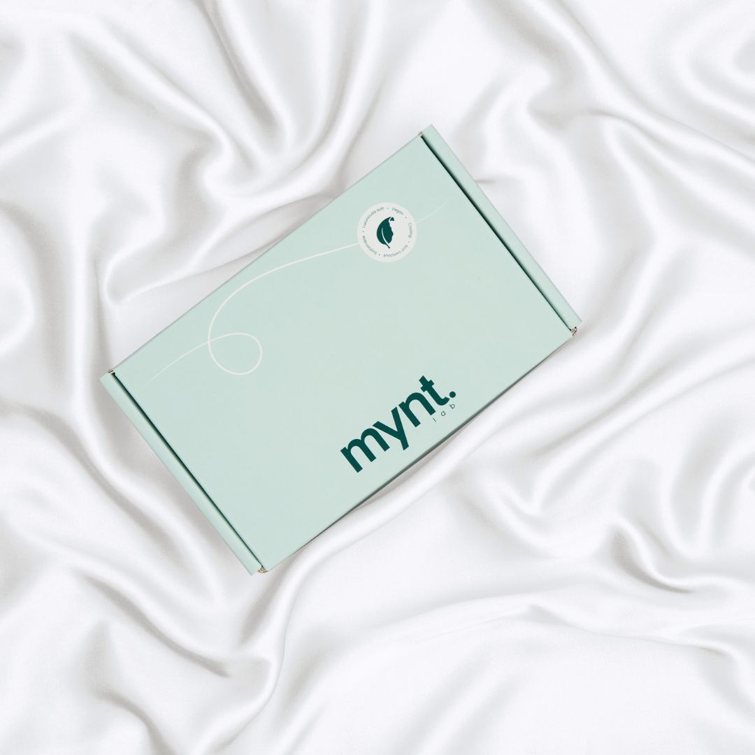 A mint green rectangular box with the mynt lab logo on the lid rests on smooth, white, silky fabric - a Pillowcase designed to enhance your beauty sleep.