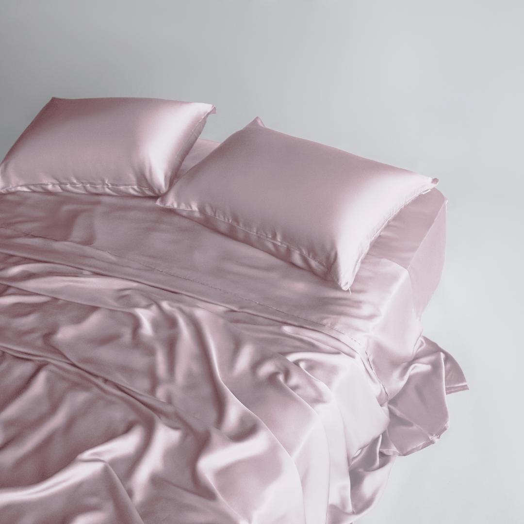 A neatly made bed features the Pink Sheet Set in light pink with a smooth, silky texture, two matching pillows, and slightly rumpled sheets near the foot, all set against a plain white background.