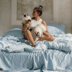 A woman sits cross-legged on a bed with the MyntFusion™ Complete Bedding Bundle from Mynt Lab, featuring light blue antibacterial cooling sheets. She holds two fluffy cats, gazing lovingly at one as sunlight softly illuminates the cozy scene.