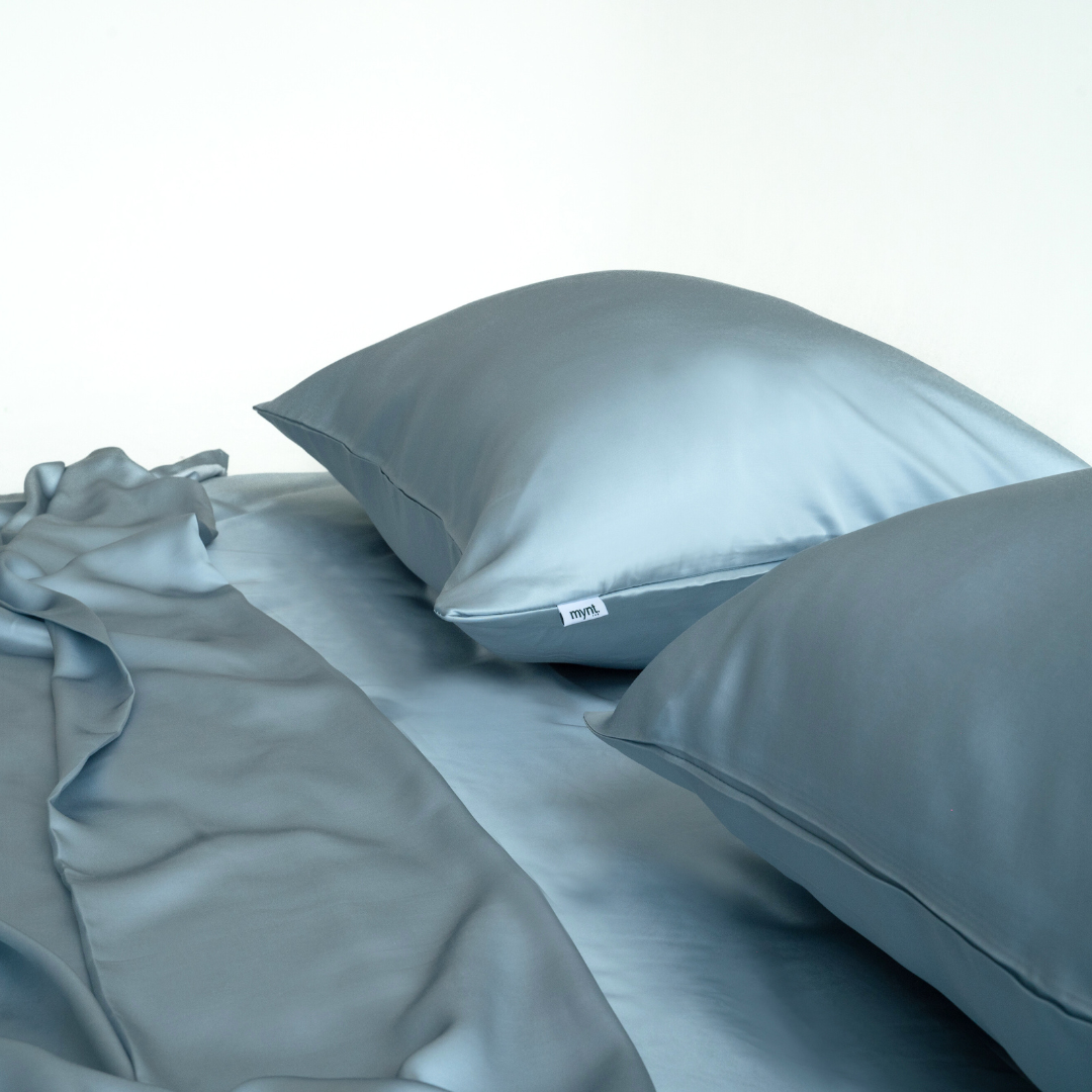 Two smooth, light blue silky pillows rest on the Powder Blue Sheet Set, their silky softness complemented by a draped fabric, creating an elegant bedroom scene.