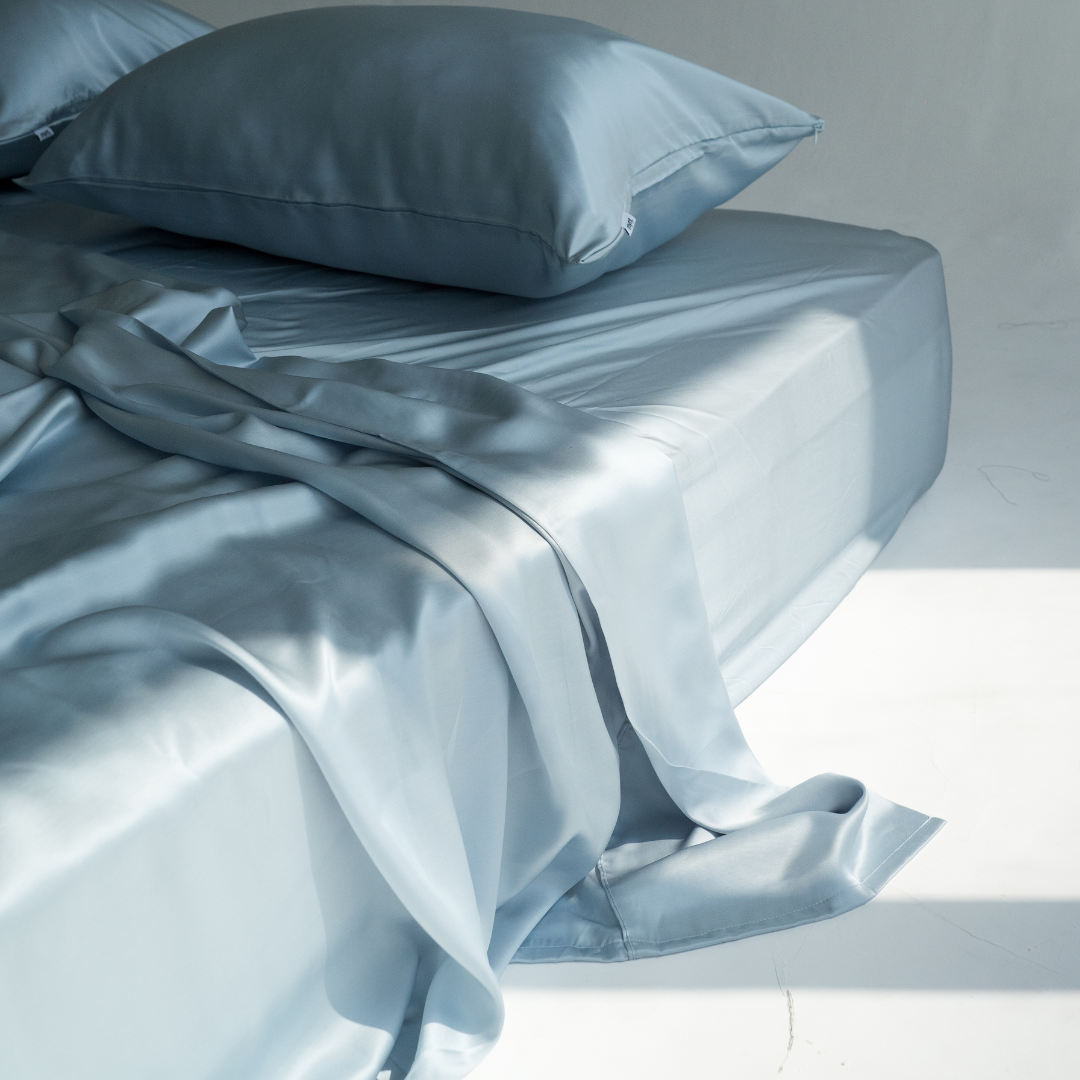 A neatly made bed with smooth, light blue satin sheets and a matching pillow. Soft sunlight casts shadows across the bedding, highlighting the fabrics silky texture.