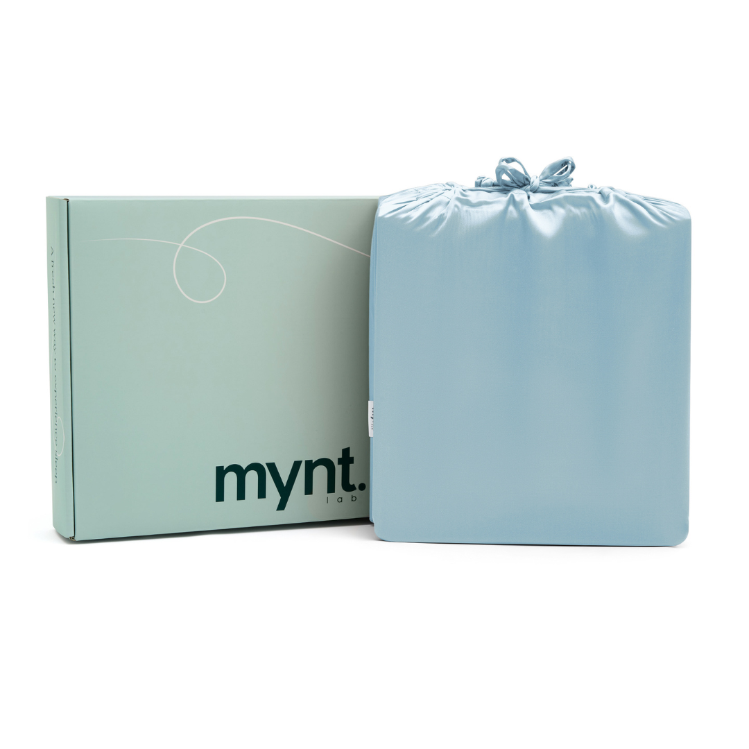 A folded light blue Fitted Sheet Set in a silky drawstring bag sits beside a mint green mynt lab-branded box.