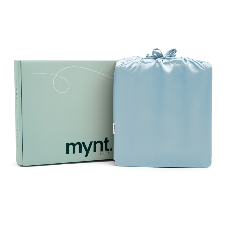 A folded light blue Fitted Sheet Set in a silky drawstring bag sits beside a mint green mynt lab-branded box.