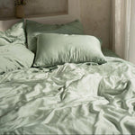 A bed dressed in light green MyntFusion™ Complete Bedding Bundle (5-Piece) by Mynt Lab is softly lit by natural light from a nearby window, with a vase of flowers placed on a recessed shelf above the pillows.