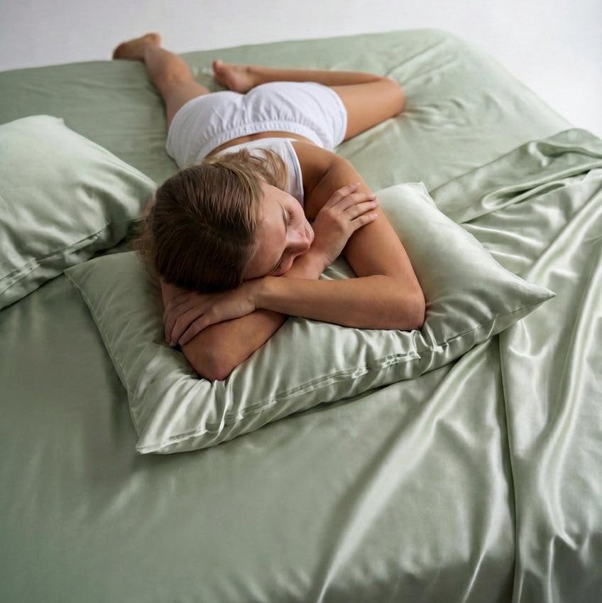 A woman in white sleepwear lies on her stomach on a bed with Mynt Lab’s MyntFusion™ Fitted Sheet Set (3-Piece) in light green satin, resting her head and arms on a matching pillow.