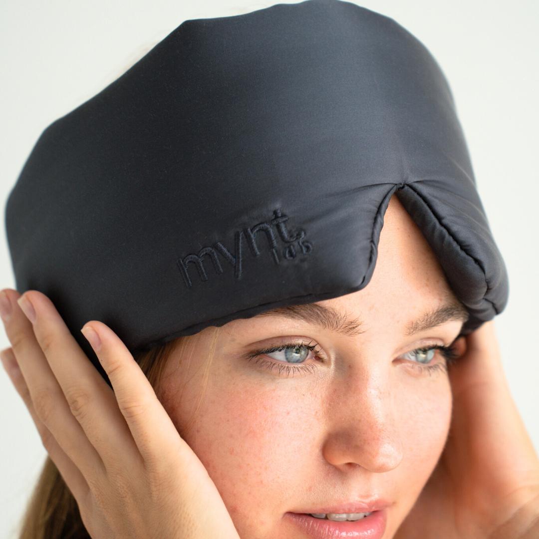 A person adjusts a charcoal, padded Sleep Mask with “mynt lab” embroidered on it, effectively blocking light from their forehead and eyes, set against a soft, neutral background.