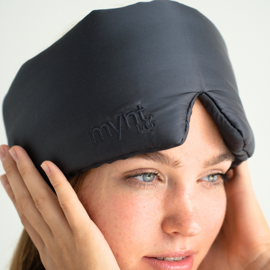 A person adjusts a dark, padded Sleep Mask with “mynt lab” embroidered on it, effectively blocking light from their forehead and eyes, set against a soft, neutral background.