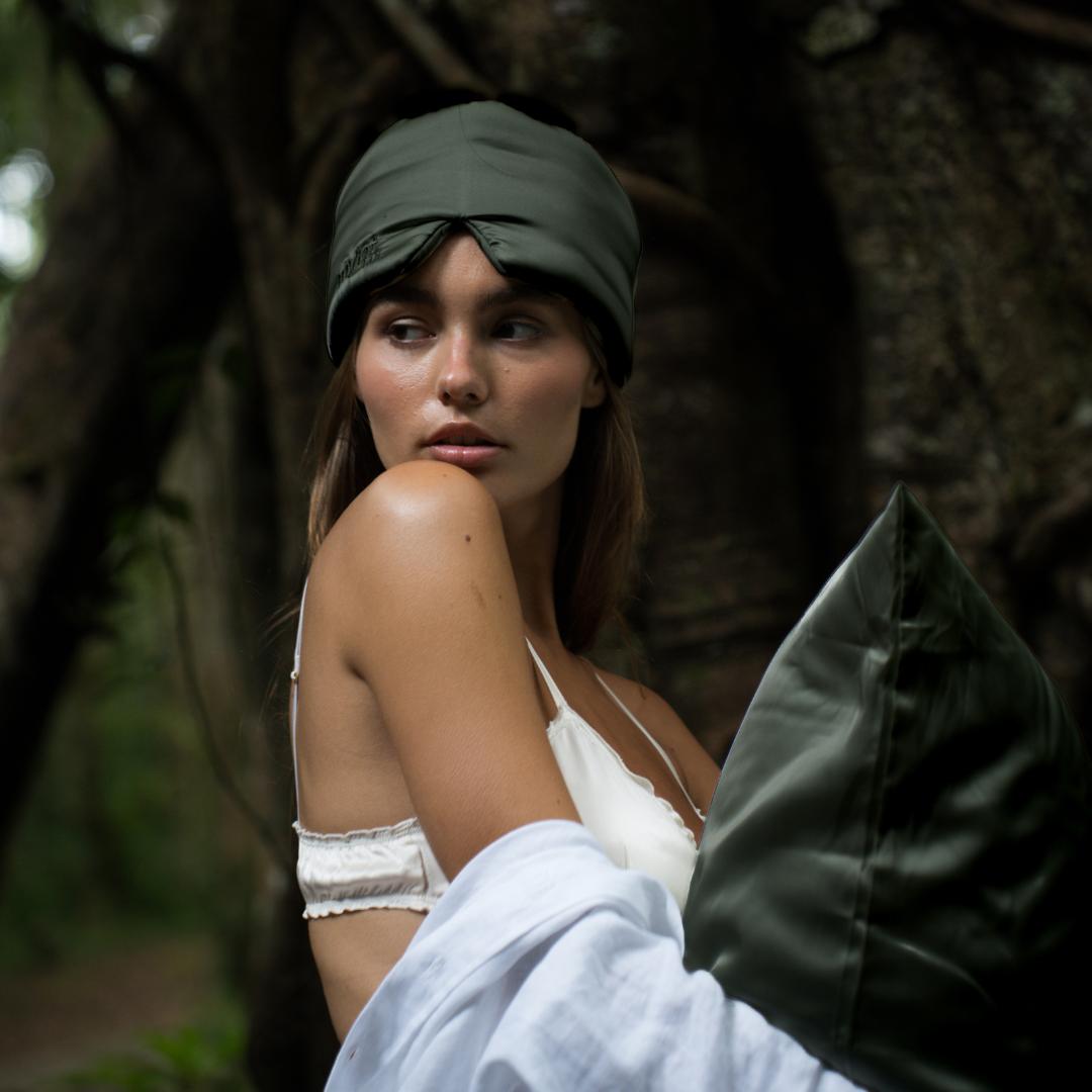 A woman in an olive green sleep mask and white top stands by a tree in the forest, holding a pillow - ideal for deep sleep. She gazes thoughtfully to the side.