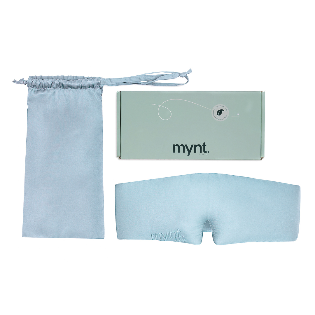 The Sleep Mask, made of light blue silky fabric, is designed to block light and promote deep sleep. It includes a matching drawstring pouch and a rectangular mynt lab branded box, all displayed on a white background.