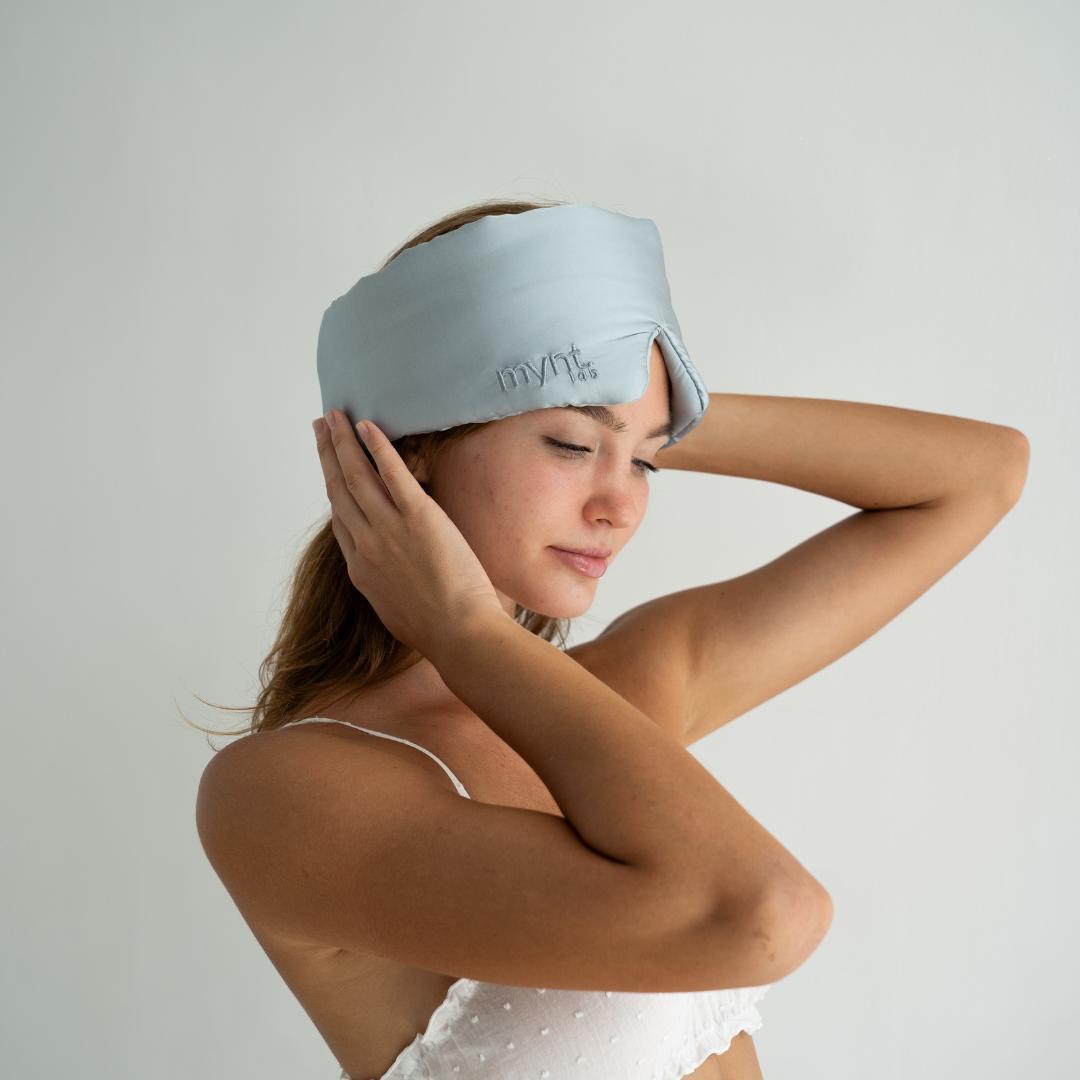 A woman with long hair wears the Sleep Mask in powder blue, covering her forehead and ears as she gently touches it. The cushioned sleep mask effectively blocks light, helping her achieve deep sleep against a plain background.