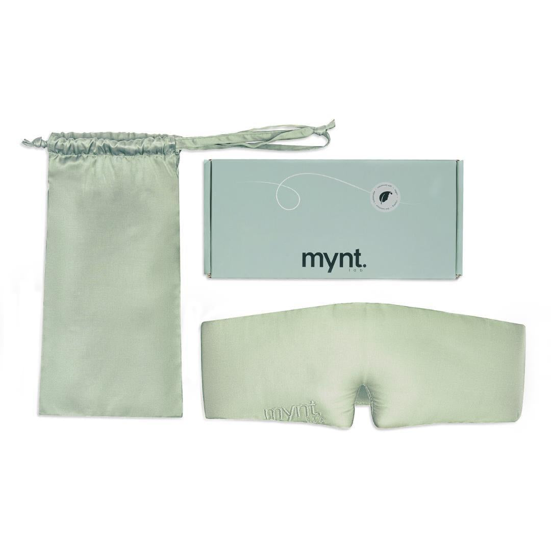 The Sleep Mask in sage green silky fabric, is ideal for blocking light and promoting deep sleep. It includes a matching drawstring pouch and comes in a rectangular mynt lab-branded box, all displayed neatly against a white background.
