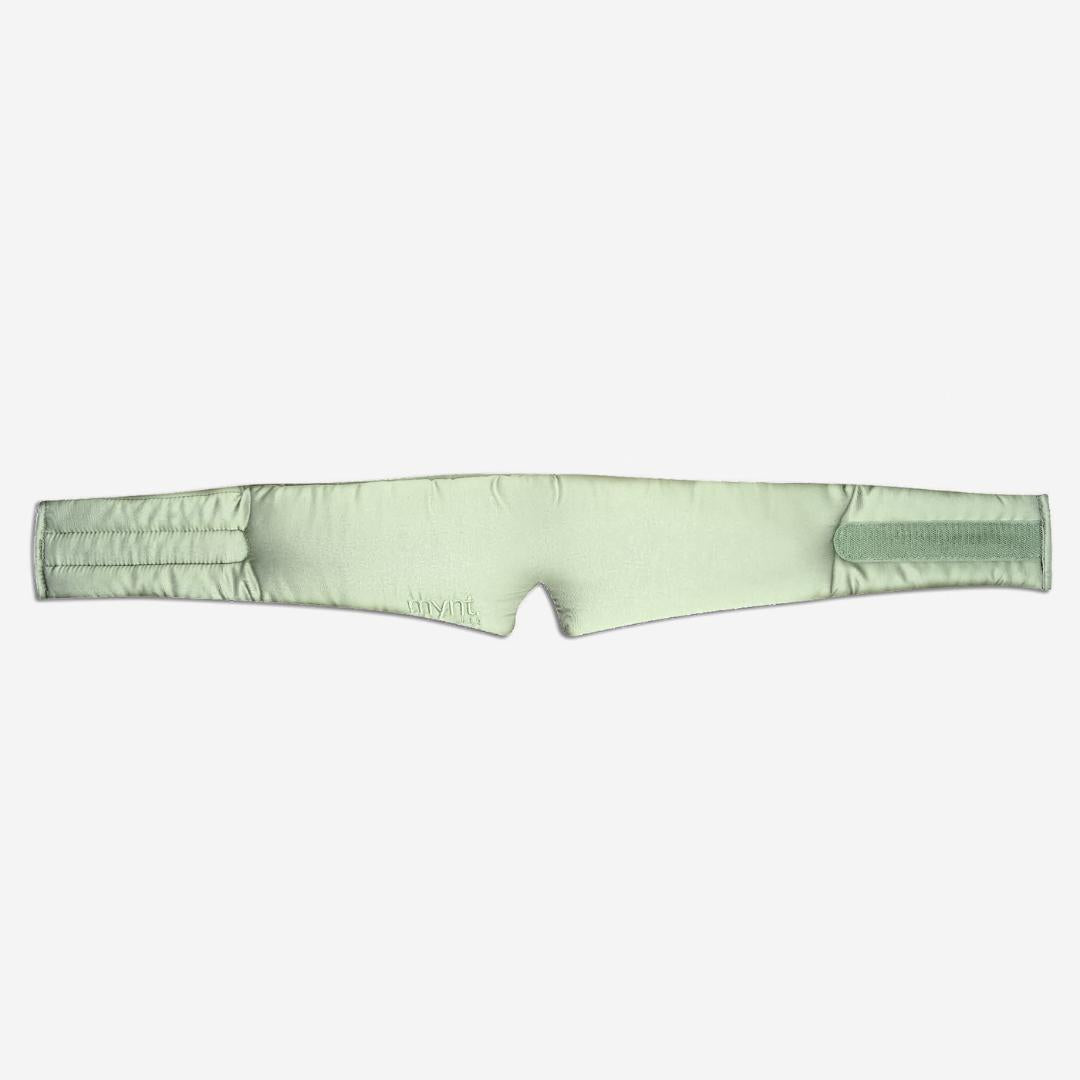 The Sleep Mask is a sage green, padded silky wrap-around design with a central notch and adjustable straps, designed to block light and promote deep sleep. Shown against a white background.