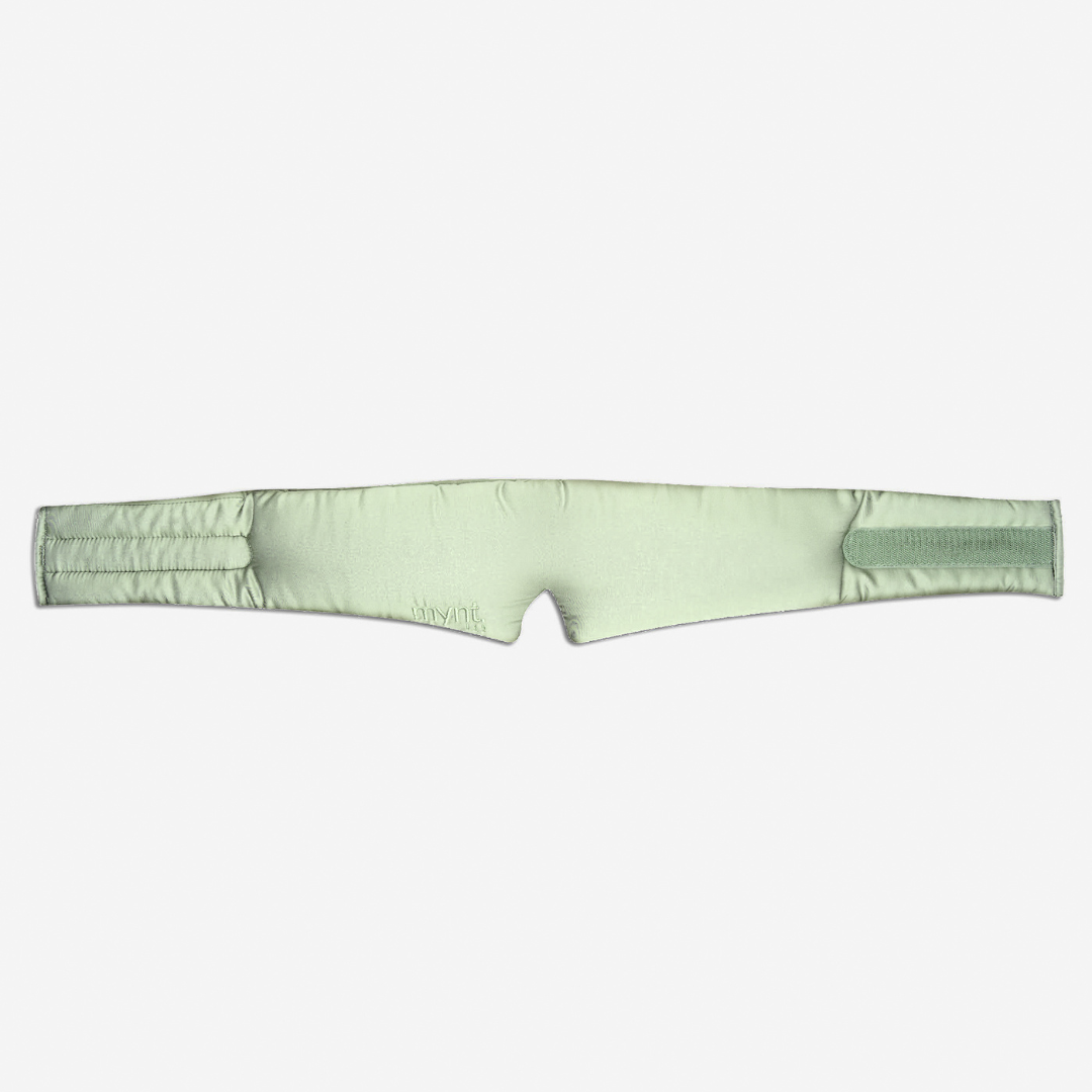The Sleep Mask is a sage green, padded silky headband with a central notch and adjustable straps, designed to block light and promote deep sleep. Shown against a white background.