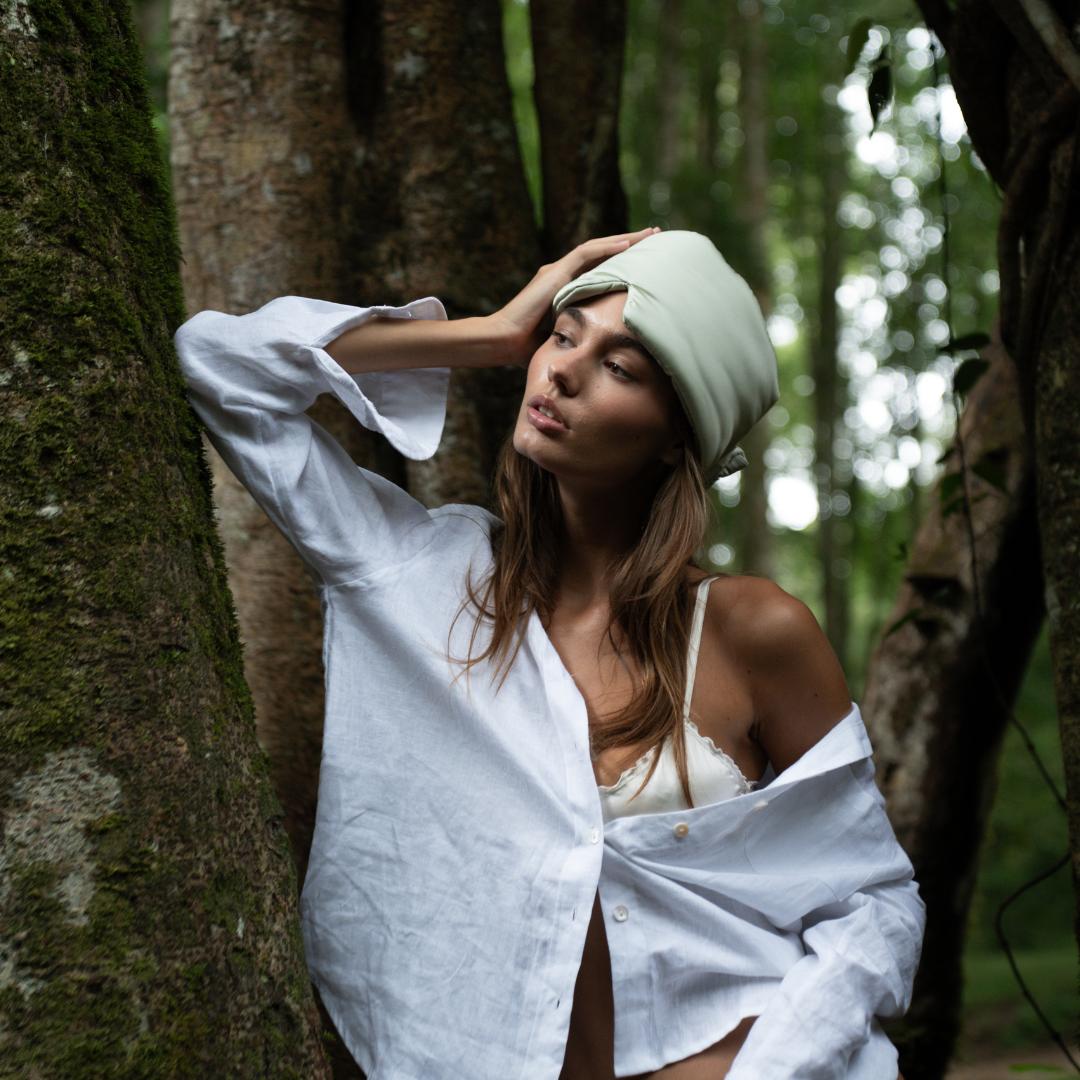 Wearing a Sleep Mask and white shirt,  a woman leans against a tree in the forest, gazing thoughtfully into the sunlit greenery behind her.