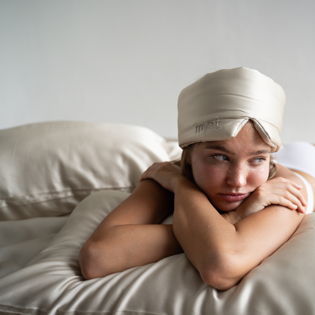 A woman rests her chin on crossed arms atop a matching silky pillow, wearing the Sleep Mask in sand, gazing to the side with a relaxed expression in a minimal, softly lit setting.