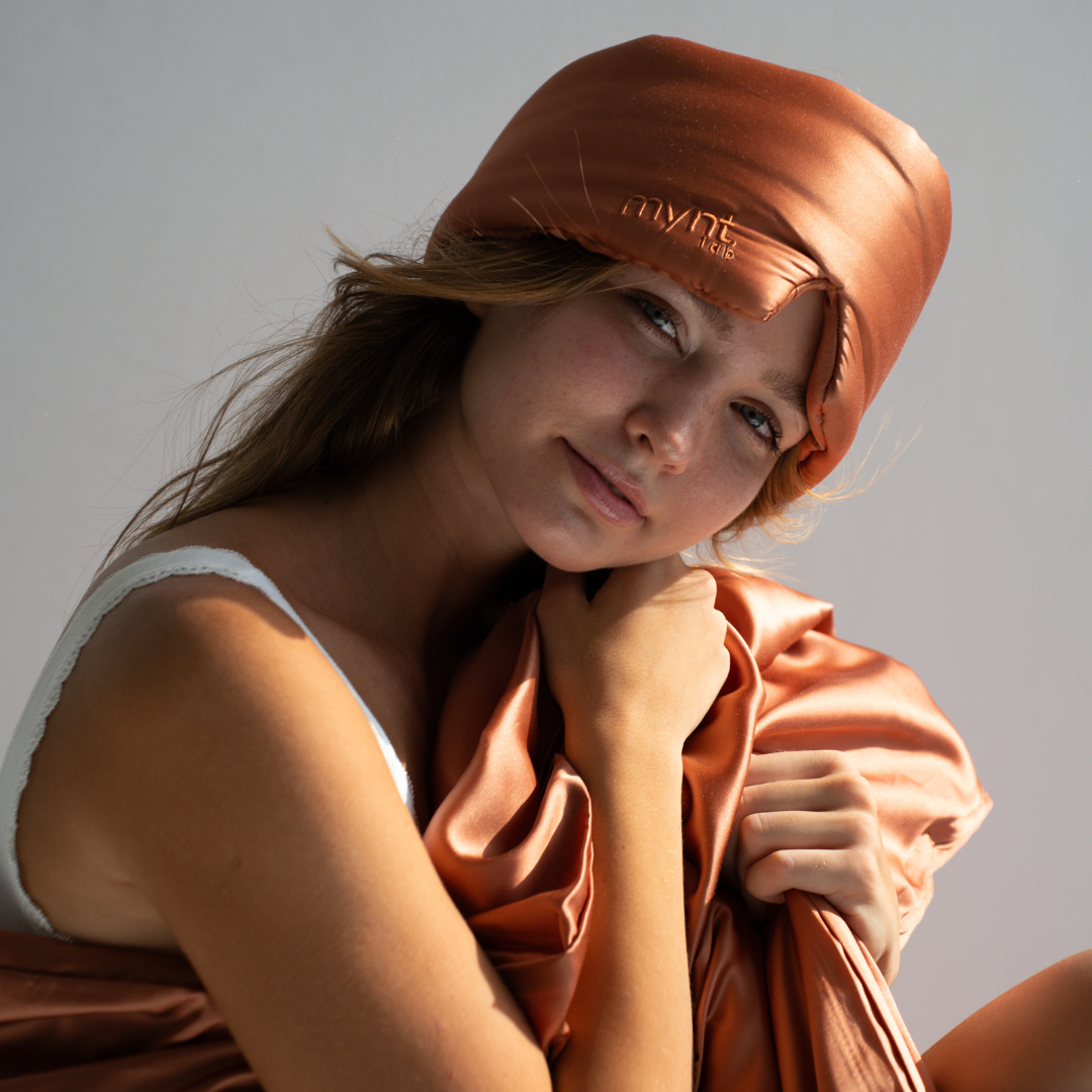 A young woman in a white tank top smiles softly at the camera, wearing the Sleep Mask and holding a matching silky sheet. The terracotta silky Sleep Mask blocks light for deep sleep as she is lit by natural sunlight.
