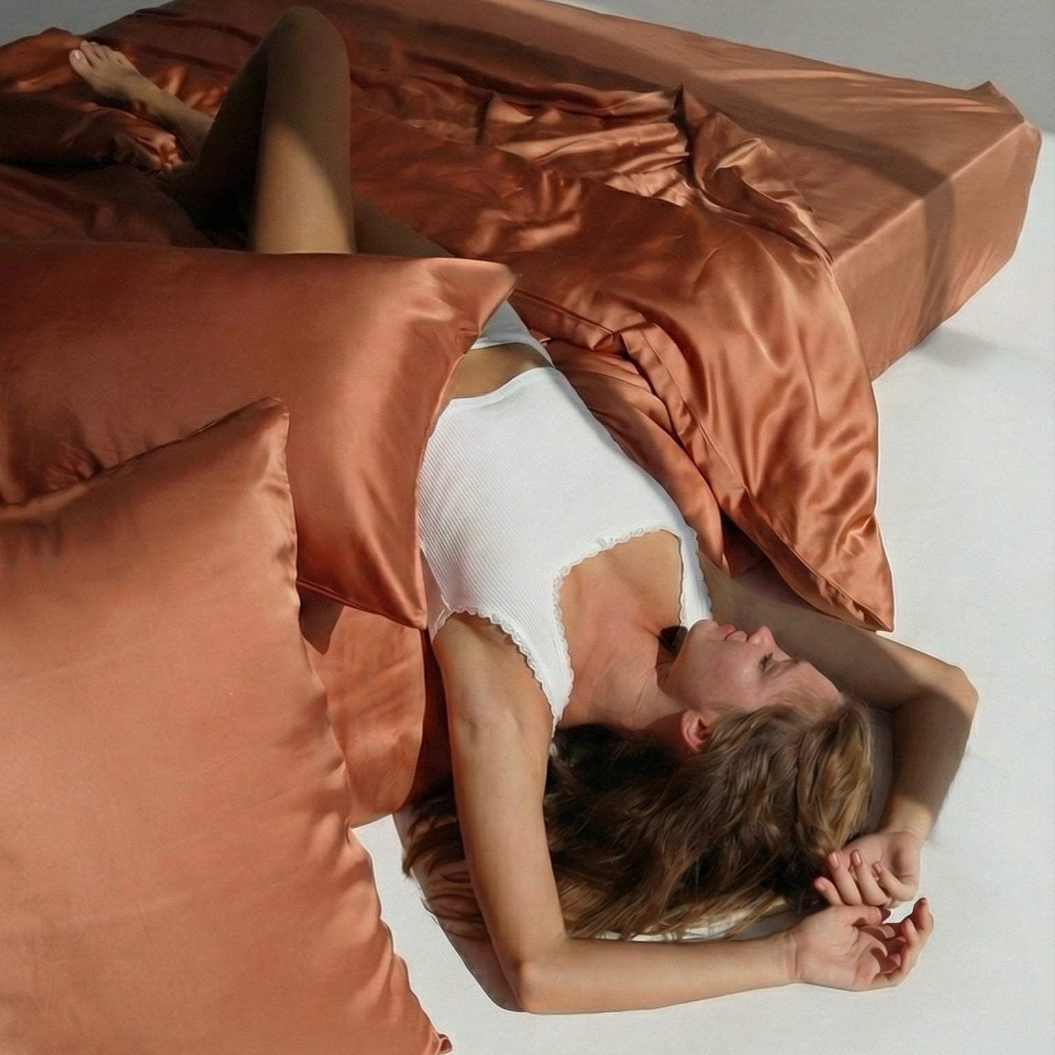 A woman in a white tank top relaxes on a bed with Mynt Lab’s MyntFusion™ Complete Sheet Set (4-Piece), featuring terracotta sheets and pillows - ideal for hot sleepers seeking cool, comfortable rest.