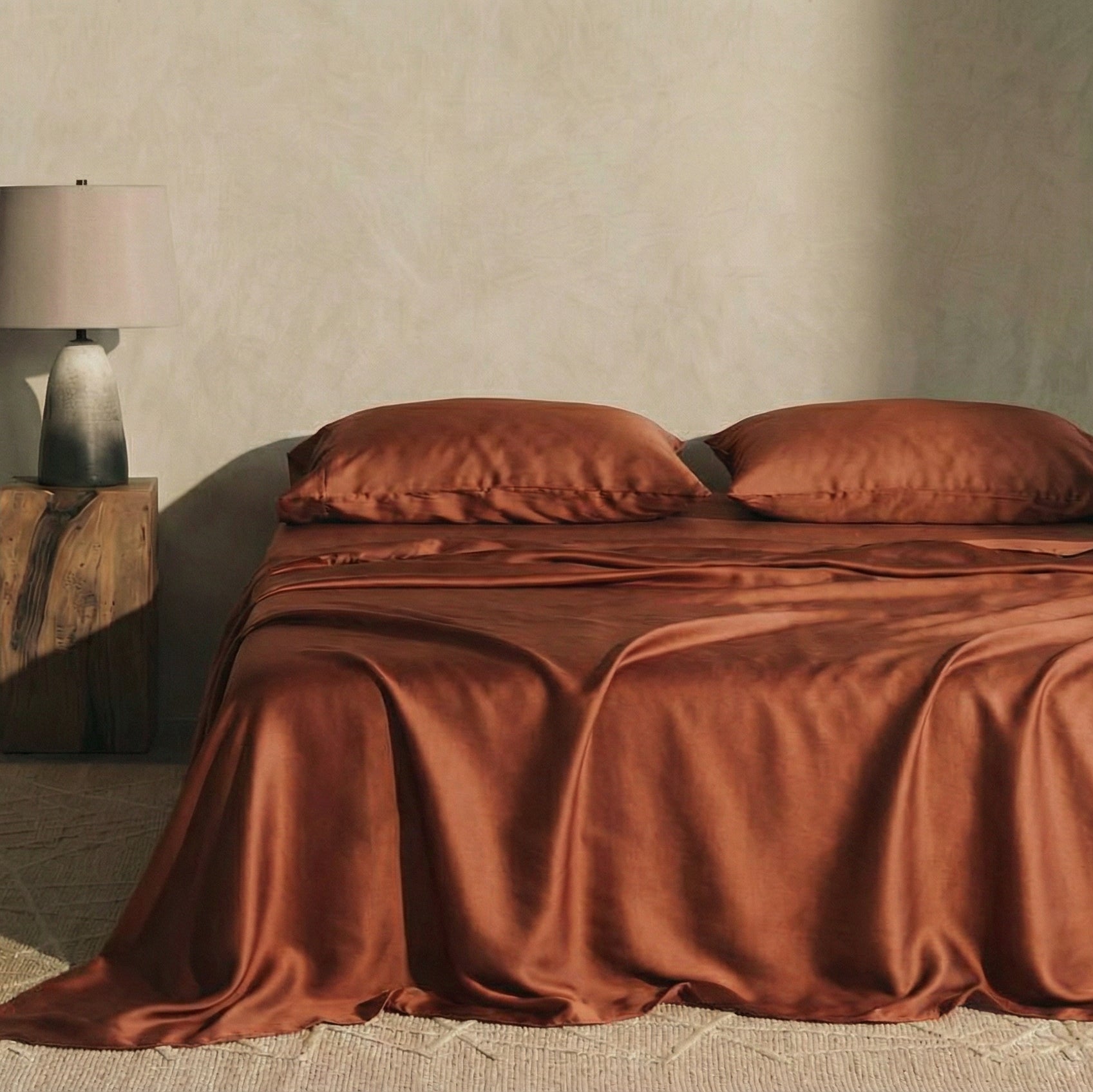 A neatly made bed with Mynt Lab’s terracotta MyntFusion™ Complete Sheet Set (4-Piece), by a wooden nightstand with a lamp - ideal for hot sleepers, set against a beige wall and carpeted floor.