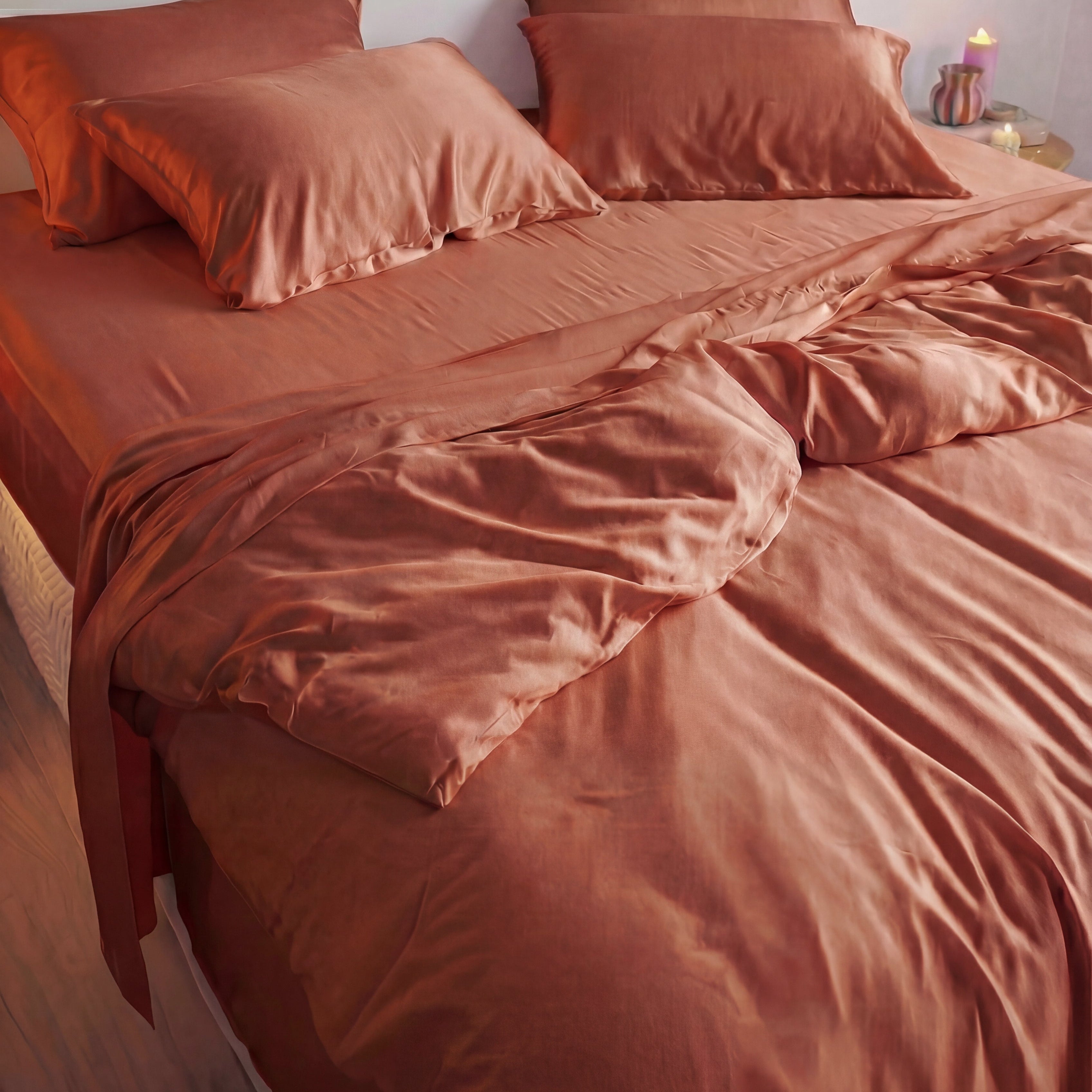 A neatly made bed with silky terracotta sheets features the Mynt Lab MyntFusion™ Duvet Cover, ideal for hot sleepers, partially turned down at the foot, with a candle on a bedside table in the background.