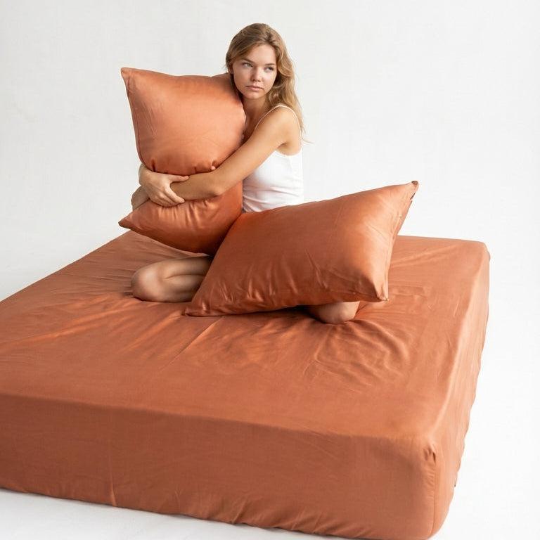 A woman in a white tank top sits on a bed made with the terracotta Mynt Lab MyntFusion™ Fitted Sheet Set (3-Piece), hugging a terracotta pillow; another pillow rests beside her against a plain white background.