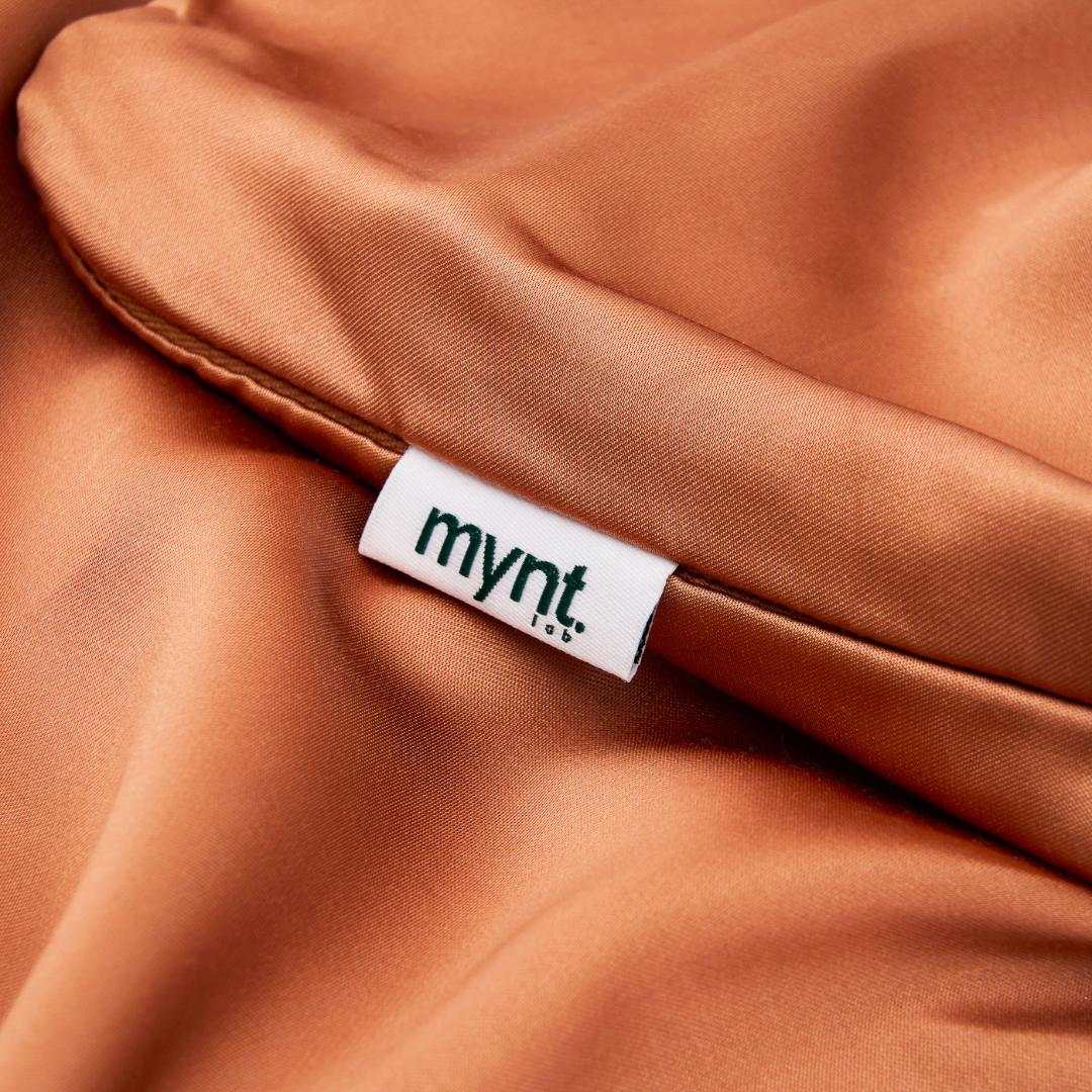 A close-up of shiny brown satin fabric with a white tag that reads mynt. in black text, sewn into the seam.