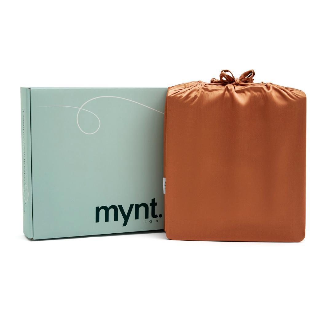 A light green box with "mynt lab" stands next to a square, terracotta-coloured silky drawstring bag - an ideal gift package for the Fitted Sheet Set or other luxurious bedding essentials.
