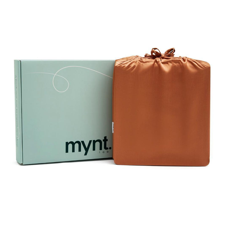 A light green box with "mynt lab" stands next to a square, terracotta-coloured silky drawstring bag - an ideal gift package for the Fitted Sheet Set or other luxurious bedding essentials.