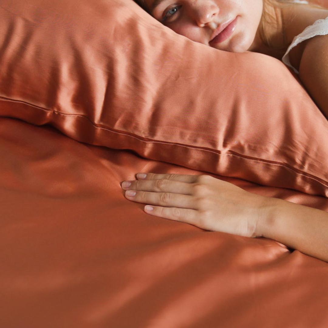 A person rests their head on a terracotta-coloured pillow with matching silky soft Fitted Sheet Set, gazing ahead with one hand gently placed on the bed.