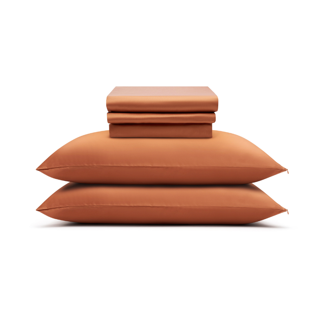Two terracotta pillows are stacked with a neatly folded Complete Sheet Set (4-Piece) placed on top, all set against a plain white background.