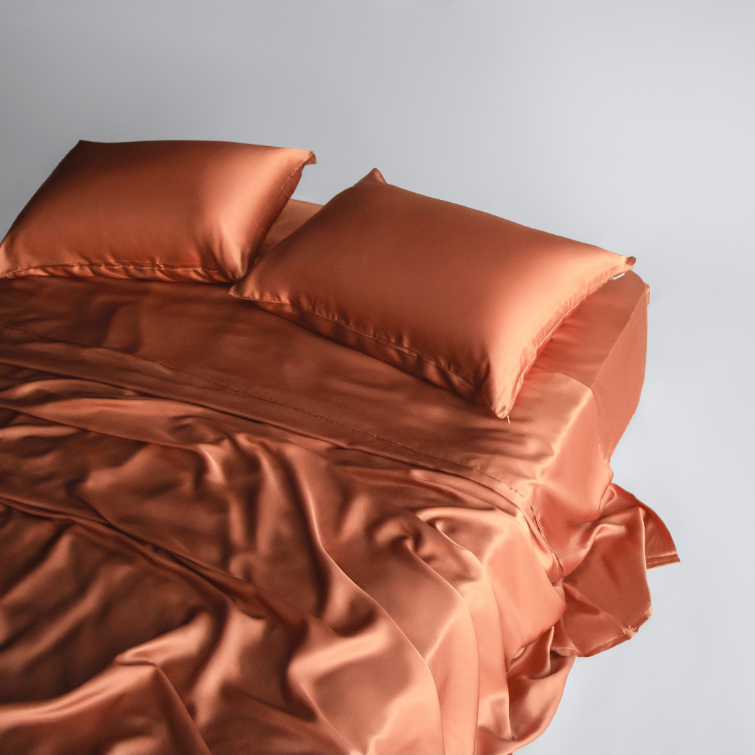 A neatly made bed featuring the Complete Sheet Set (4-Piece) in terracotta, with silky soft fabric and matching pillows, photographed from above on a plain light grey background. The smooth, shiny sheets drape elegantly with gentle folds.