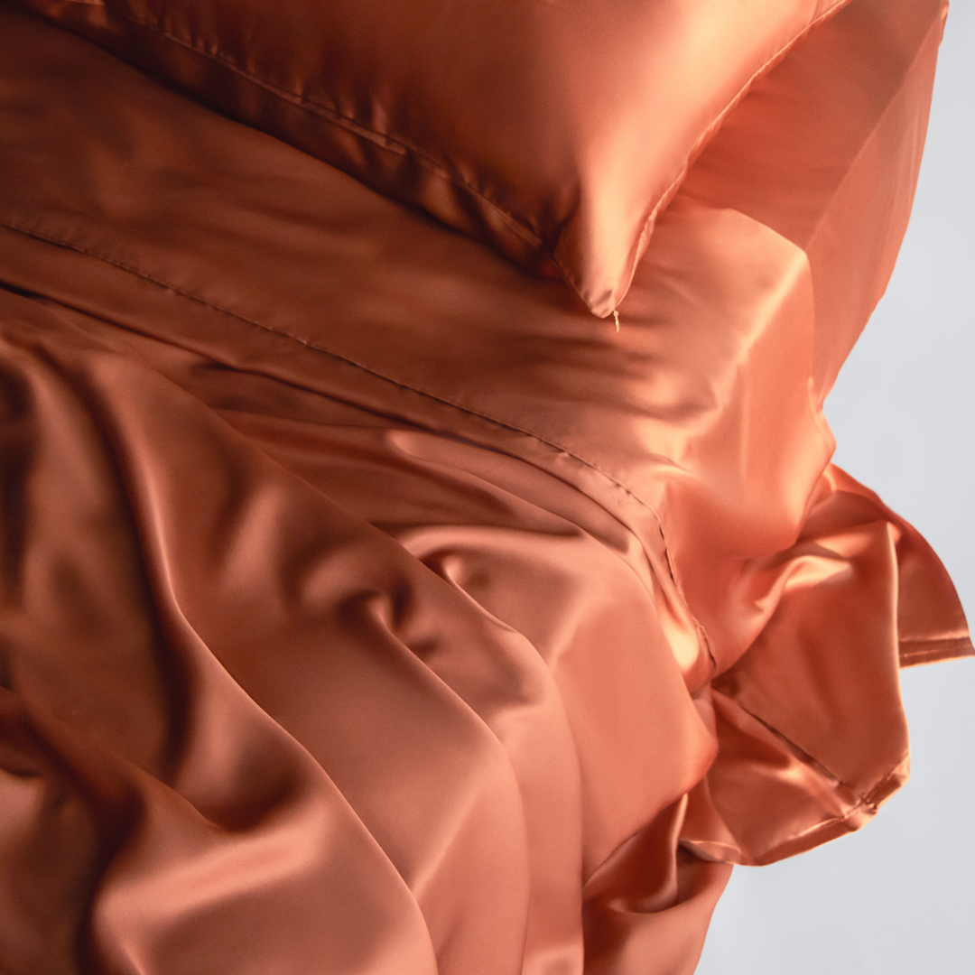 A close-up of a bed dressed in the Complete Sheet Set (4-Piece) features shiny, terracotta silky soft sheets and a matching pillowcase, highlighting the smooth folds and luxurious, soft reflective texture.