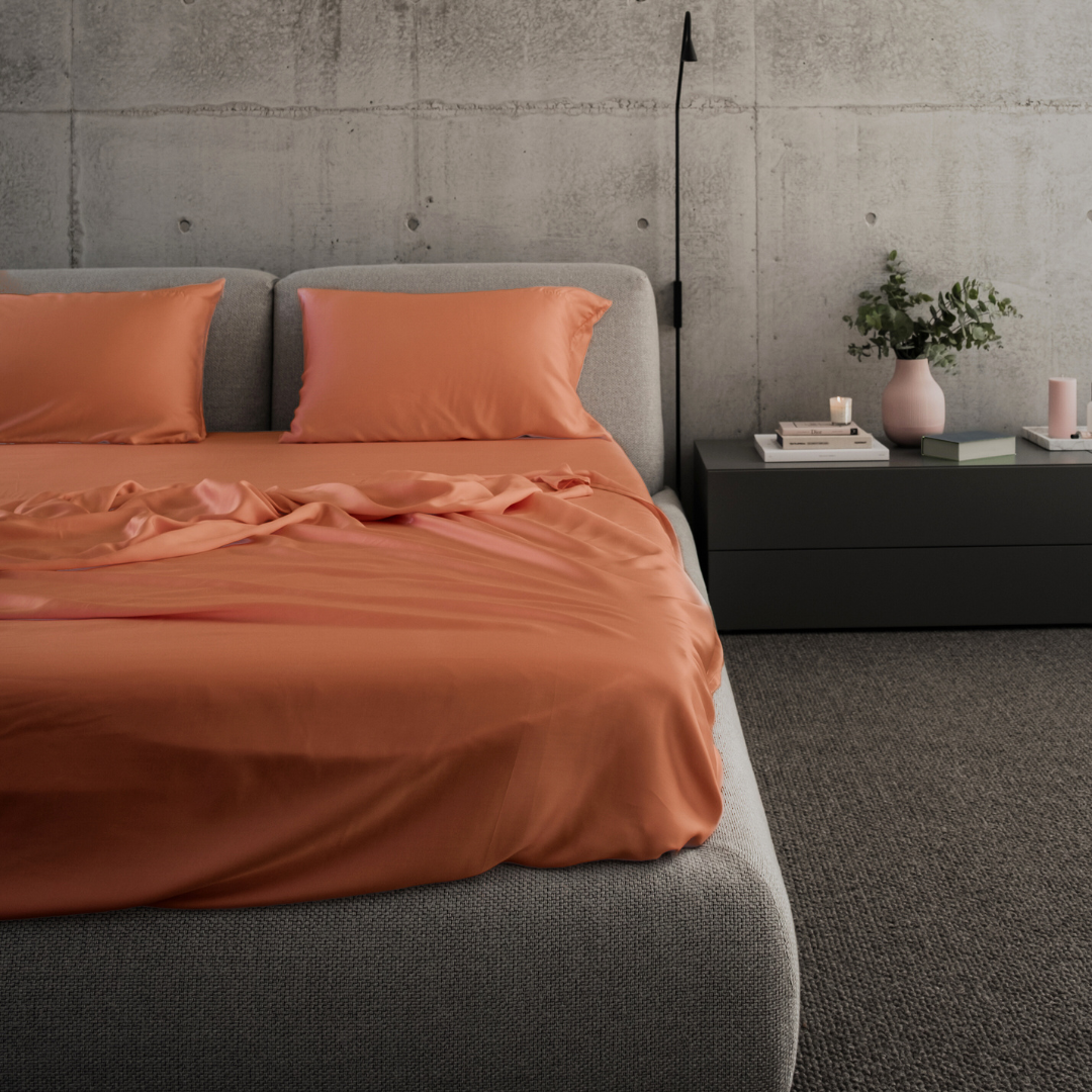 A modern bedroom with a grey upholstered bed featuring orange sheets and pillows, a dark bedside table with a pink vase, books, and a candle, and a concrete wall in the background.